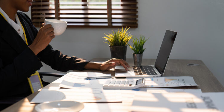 A imagem mostra uma pessoa sentada em uma mesa com um laptop aberto à sua frente. A pessoa está segurando uma xícara em uma mão e parece estar usando o laptop com a outra. Na mesa, há vários papéis espalhados, alguns dos quais contêm gráficos e tabelas. Também há um prato vazio e uma pequena planta em vaso ao lado do laptop. A sala recebe luz natural através das persianas de uma janela, sugerindo um ambiente de trabalho em casa ou no escritório.
