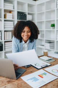 A imagem mostra uma pessoa sentada em uma mesa com um laptop aberto à sua frente. A pessoa está segurando e olhando alguns papéis. Na mesa, há mais papéis com gráficos e tabelas, sugerindo que a pessoa pode estar analisando dados ou revisando relatórios. Ao fundo, há uma estante cheia de livros, contribuindo para a impressão de um ambiente de escritório ou estudo. O rosto da pessoa está obscurecido por um retângulo marrom, impedindo a identificação. 